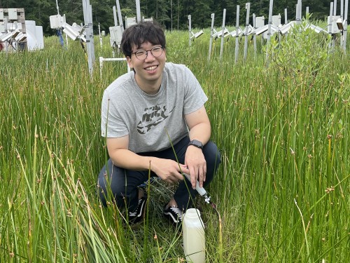 Man wearing glasses ans short sleeve shirt crouches down for a photo in a field of tall green grass.