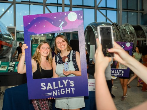 Two women pose with a large paper cutout as someone uses their cellphone to take a picture